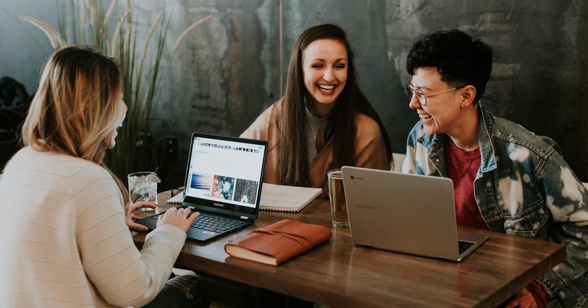 Trois personnes rient ensemble autour d'une table de café, chacune devant son ordinateur portable.